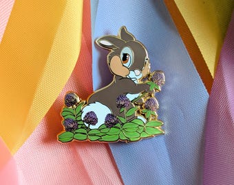 Peut inclure: Épingle en émail doré représentant un lapin de dessin animé avec une fourrure marron, un ventre blanc et de grands yeux. Le lapin est entouré de feuilles vertes et de fleurs violettes. L'épingle est posée sur un fond de rubans colorés.