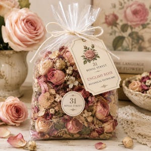 May include: A clear plastic bag filled with dried botanical potpourri, featuring pink and cream-colored rosebuds and small white flowers. A cream-colored tag reads 31 ROYAL STREET ENGLISH ROSE BOTANICAL POTPOURRI, tied with twine.