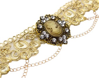 May include: Gold chain bracelet with a delicate lace design and a large, ornate cameo pendant featuring a woman's profile. The pendant is set with clear rhinestones.