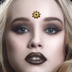 May include: A close-up of a woman's face with dark eye makeup and dark brown lipstick. She is wearing a small, round, gold and black jeweled sticker on her forehead.