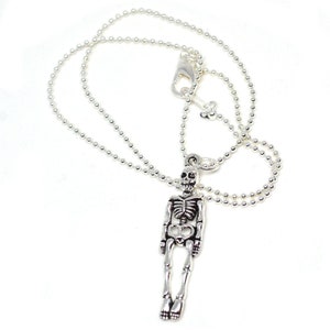 May include: Silver-toned skeleton pendant necklace. The pendant is a detailed miniature skeleton with articulated limbs, suspended from a delicate ball chain. The necklace has a lobster clasp closure. The background is white.