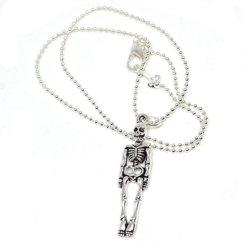 May include: Silver-toned skeleton pendant necklace. The pendant is a detailed miniature skeleton with articulated limbs, suspended from a delicate ball chain. The necklace has a lobster clasp closure. The background is white.