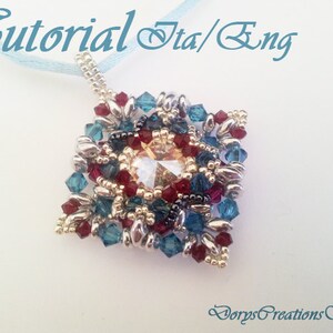 Può includere: Un ciondolo a forma quadrata composto da perline, con un centro in cristallo, che presenta un mix di perline argentate, dorate, blu e rosse. Il ciondolo è attaccato a un nastro blu.