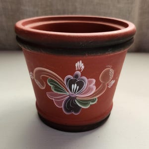 以下が含まれることがあります： 手描きの花柄が施されたテラコッタ色のセラミック植木鉢。 植木鉢は濃い茶色の縁と底があり、花柄にはピンク、紫、緑、白のアクセントが含まれています。 この植木鉢は小さな植物に適しています。