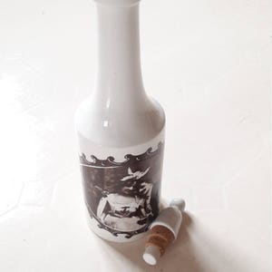以下が含まれることがあります： コルク栓が付いた白いセラミックボトル。ボトルには、ドレスを着た女性の白黒写真が描かれています。