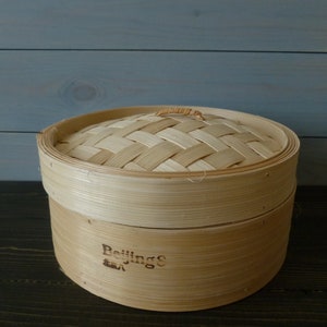 May include: A round, natural bamboo steamer with a woven lid. The steamer has a brown label that reads "Beijing's" with a small symbol below it.