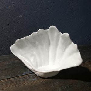 May include: White ceramic bowl shaped like a seashell. The bowl has a smooth, glossy finish and a slightly ruffled edge.