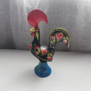 Puede incluir: Una figurita de gallo negro con detalles pintados en rojo, amarillo y verde. El gallo está de pie sobre una base azul con la palabra "Portugal" escrita en ella.