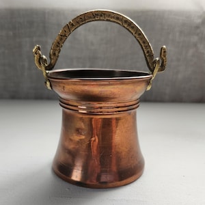 Pode incluir: Um pequeno balde de cobre com base alargada e borda larga. O recipiente possui uma alça de latão curva e texturizada, fixada nas laterais com alças de metal. A superfície de cobre tem um acabamento metálico polido com variações de tom.