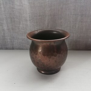 Pode incluir: Um pequeno vaso de cobre martelado com base arredondada. O vaso tem uma pátina marrom escura e é perfeito para exibir plantas pequenas ou suculentas.
