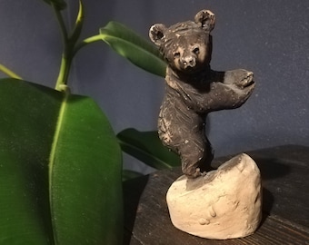 May include: A small ceramic bear figurine standing on a rock base. The bear is brown and has a textured surface. The rock base is a light brown color.