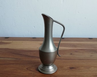 May include: A silver pewter vase with a handle, standing on a wooden surface.
