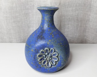 May include: A blue ceramic vase with a textured surface and a floral design. The vase has a rounded body, a narrow neck, and a small opening. The floral design is in the centre of the vase.