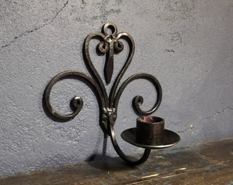 Puede incluir: Candelabro de pared de metal negro con un diseño decorativo de volutas. El candelabro tiene una sola copa para velas y está montado en una superficie de madera oscura.