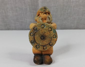 May include: A ceramic figurine of a warrior holding a shield. The figurine is brown and green with a textured surface. The warrior is wearing a helmet and has a beard.