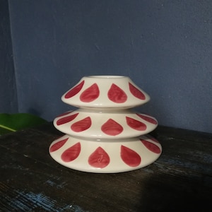 May include: A white ceramic candle holder with three tiers. Each tier is decorated with a pattern of red teardrop shapes.