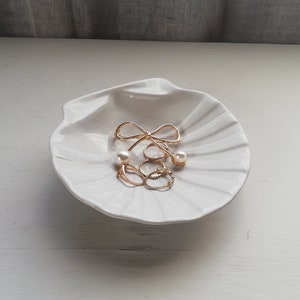 Może przedstawiać: Biała ceramiczna miseczka w kształcie muszli z złotą kokardką i perłowymi kolczykami i pierścionkami.