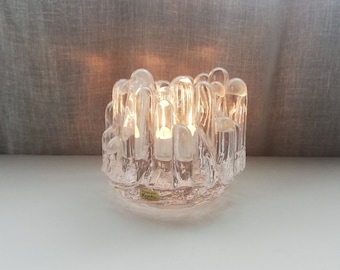 May include: A clear glass candle holder with a unique, textured design. The holder features multiple vertical glass tubes that create a modern and stylish look.