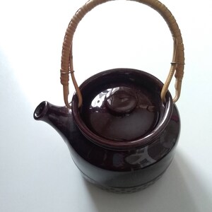 May include: A brown ceramic teapot with a woven bamboo handle. The teapot has a lid and a spout.