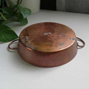 May include: A copper-coloured, round cooking pot with two small handles. The pot has a slightly distressed appearance with areas of discolouration. The pot is sitting on a white surface. The pot is likely used for cooking.