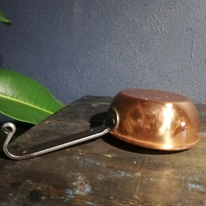 May include: A copper saucepan with a black metal handle. The handle has a hook on the end.