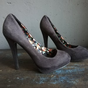 Op de afbeelding: Een paar zwarte su&egrave;de pumps met een bloemenprint voering. De hakken zijn ongeveer 10 cm hoog.