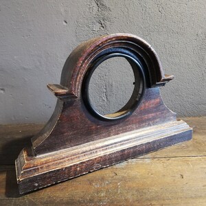 May include: A dark brown wooden clock face with a black metal ring around the center. The clock face is shaped like an arch and has a decorative design on the top and bottom.