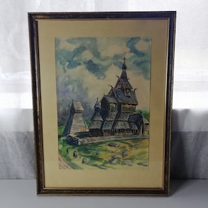 Puede incluir: Una pintura al agua enmarcada que representa una iglesia de madera con un techo empinado y una alta aguja. La iglesia está pintada en tonos de marrón y gris, con una zona verde de hierba en primer plano. El cielo es de un azul claro con nubes blancas.