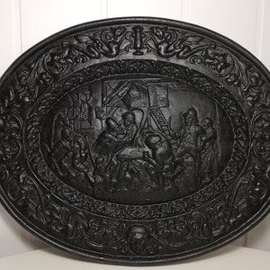 May include: Black cast iron wall plaque with an oval shape and a raised design of a scene with people gathered around a table. The plaque is decorated with intricate floral patterns around the border.