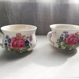 Op de afbeelding: Twee witte keramische mokken met bloemenmotieven. De mokken hebben een brede, afgeronde vorm en een enkele handgreep. Het bloemenmotief heeft roze rozen, paarse en blauwe bloemen en groene bladeren.