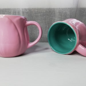 May include: Two ceramic mugs, one pink with a light green interior and one light green with a pink interior. Both mugs have a rounded shape and a handle.