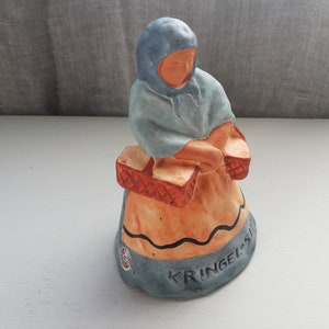 Può includere: Una statuetta in ceramica di una donna che indossa un mantello blu con cappuccio e porta due cesti. La statuetta è dipinta in tonalità di blu, arancione e marrone. La base della statuetta ha il testo "KRINGEL-S" scritto su di essa.