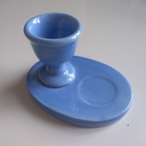 May include: A light blue ceramic egg cup with a matching saucer. The egg cup has a rounded base and a wide, shallow top. The saucer has a small indentation for the egg cup to sit in.