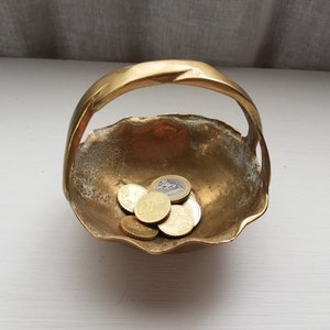 May include: A small, gold-colored brass bowl with a handle. The bowl is shaped like a shell and has a scalloped edge. There are several coins inside the bowl.