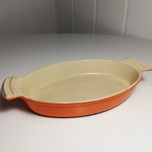 May include: Orange oval-shaped baking dish with two handles. The dish is made of enamel-coated cast iron and has a cream-colored interior.