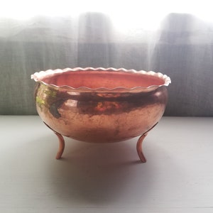 Op de afbeelding: Een koperen plantenbak met een gegolfde rand en drie poten. De plantenbak is gemaakt van gehamerd koper en heeft een rustieke, vintage uitstraling.