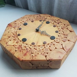 May include: A wooden clock with an octagonal design. The clock face is light wood, featuring black and brown markers. The frame is constructed from small, circular wooden pieces, giving a rustic appearance. The clock hands are black. The clock measures approximately 30 cm across.