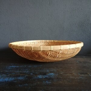 May include: A large, round, woven basket made of natural materials. The basket is light brown in color and has a smooth, even texture. It is sitting on a dark wooden surface.