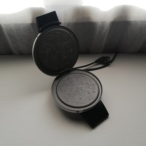 May include: A black waffle maker with a decorative pattern on the cooking plates. The waffle maker is open and the plates are facing up.