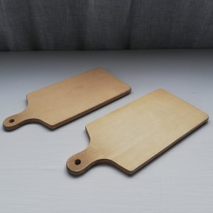 Puede incluir: Dos tablas de cortar de madera con asas. Las tablas son de color marrón claro y tienen una superficie lisa.