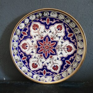 May include: A round decorative plate with a gold rim and a white background. The plate features a blue and red floral design with intricate details.