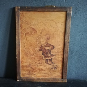 May include: A framed wood-burned artwork depicting a girl in a winter scene. The girl is wearing a hat and coat and is holding a stick. The background is a forest with trees and snow.