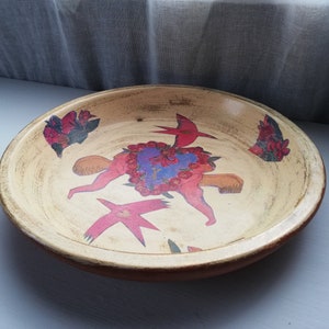 May include: A round ceramic serving bowl with a cream-colored background and a colorful painted design. The design features two figures with red and pink wings, a heart with flowers, and floral accents around the edge.