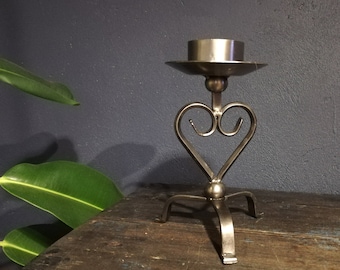 May include: A silver metal candle holder with a heart-shaped design and three legs. The holder is made of wrought iron and has a rustic finish.