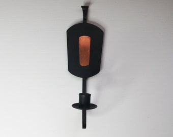 Puede incluir: Aplique de pared de metal negro con un recorte rectangular y un portavelas. El recorte tiene un fondo de color cobre.