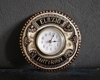 May include: A round, copper-coloured wall clock with intricate decorative details. The clock face is white with black numbers and hands. The clock is inscribed with the words "ELAZIG HATIRASI".