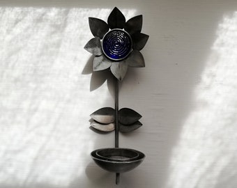 Peut inclure: Une décoration murale en métal en forme de tournesol avec un centre en verre bleu et un petit bol en bas. Le tournesol est fixé à une tige en métal.