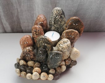 May include: A candle holder made of various sized and colored stones with painted eyes. The stones are arranged in a circular shape with a small tea light candle in the center.