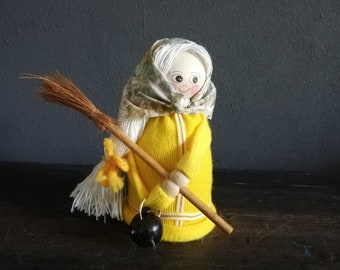 May include: A handmade witch doll with white yarn hair, a yellow knitted dress, a floral patterned headscarf, and a brown broom.