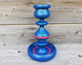 May include: A blue and pink wooden candle holder with a round base and three tiers. The top tier is a cup-shaped holder, the middle tier is a round ball, and the bottom tier is a wide, flat base.